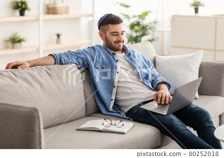 Cheerful male jew working on laptop at home on sofa 80252018
