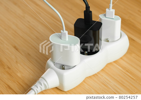 The electrical extension strip with connected white and black power plugs on the wooden table. Smart phones chargers in power strip for electrical sockets 80254257