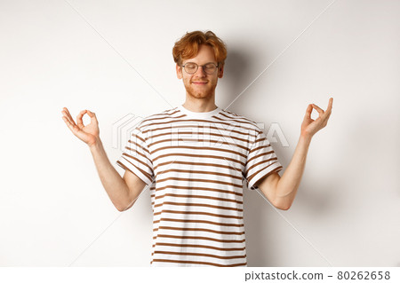Calm and relaxed young man with red messy hair, spread hands sideways in mudra gesture and smiling, practice yoga or meditating, white background 80262658
