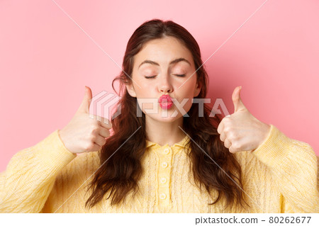 Beauty and fashion. Close up portrait of lovely and coy girl pucker lips with eyes closed, showing thumbs up in approval, praise something good, standing against pink background 80262677