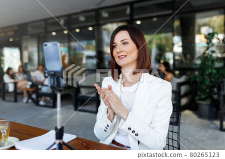 Modern smartphone gadget,tripod record young female speaker,coach making live broadcast Internet, cellphone shoot woman blogger or influencer talking for tutorial or webinar online. street cafe place. 80265123