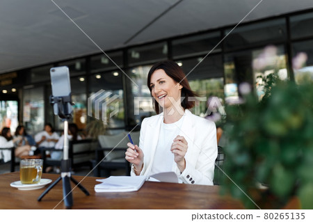 Modern smartphone gadget,tripod record young female speaker,coach making live broadcast Internet, cellphone shoot woman blogger or influencer talking for tutorial or webinar online. street cafe place. 80265135