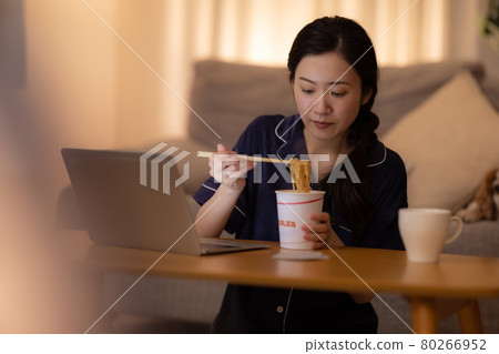 Living room at night A woman eating cup ramen while looking at a computer 80266952