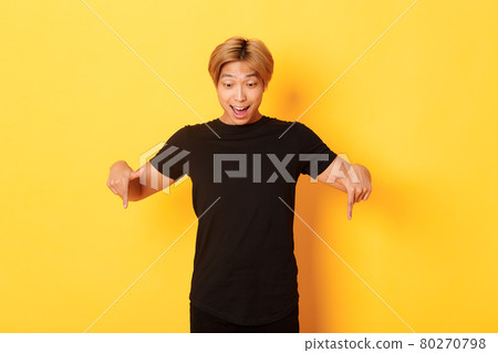 Portrait of excited and happy asian guy with blond haircut, pointing and looking down amused, standing yellow background 80270798