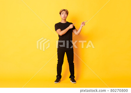 Full-length of sad and miserable asian guy in black clothes, pouting, pointing fingers upper right corner, standing yellow background Full-length of sad and miserable asian guy in black clothes, pouting, pointing fingers upper right corner, standing yellow background 80270800