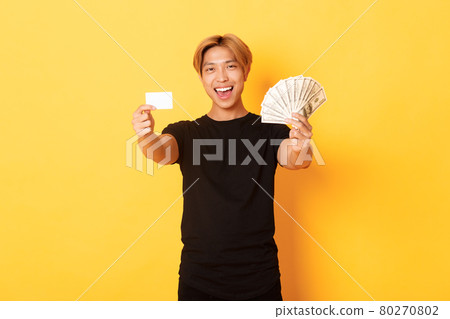 Happy good-looking asian guy in black casual clothes, showing money and credit card, smiling sassy, yellow background Happy good-looking asian guy in black casual clothes, showing money and credit card, smiling sassy, yellow background 80270802