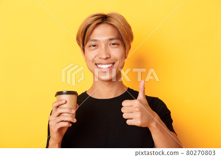 Close-up of satisfied asian handsome guy recommend cafe, holding cup of coffee and showing thumbs-up in approval, smiling pleased over yellow background Close-up of satisfied asian handsome guy recommend cafe, holding cup of coffee and showing thumbs-up in approval, smiling pleased over yellow background 80270803