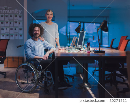 Businessman in a wheelchair in modern coworking office space working late night in office. Colleagues in background. Disability and handicap concept. Businessman in a wheelchair in modern coworking office space working late night in office. Colleagues in background. Disability and handicap concept. 80271311
