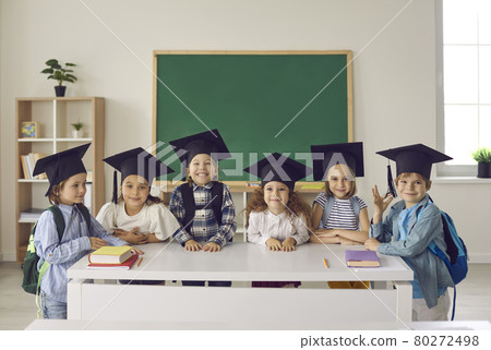 Group of happy school kids in academic caps standing by table with books in classroom 80272498