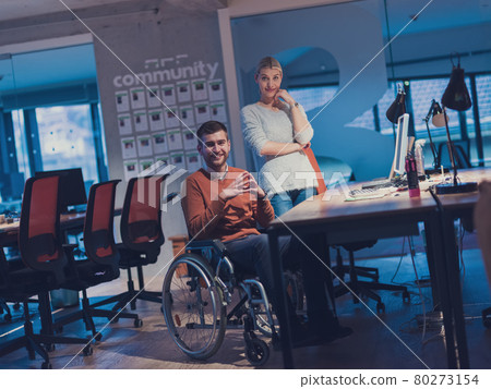 Businessman in a wheelchair in modern coworking office space working late night in office. Colleagues in background. Disability and handicap concept.  80273154