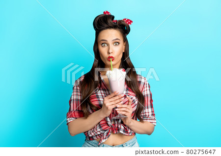 Shocked young pinup woman in retro clothes drinking yummy milk shake on blue studio background Shocked young pinup woman in retro clothes drinking yummy milk shake on blue studio background 80275647