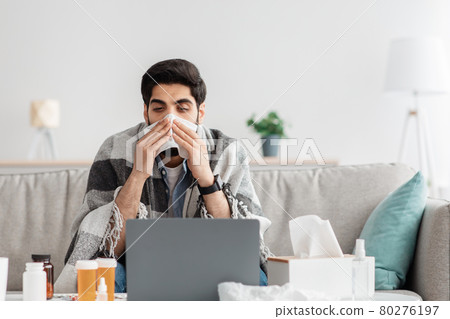 Sick arab man sneezing nose in paper tissue and looking at laptop screen, sitting at home and working, empty space 80276197