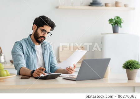 Paying bills, taxes at home online. Young arab man using calculator and laptop computer, sitting at kitchen, copy space Paying bills, taxes at home online. Young arab man using calculator and laptop computer, sitting at kitchen, copy space 80276365