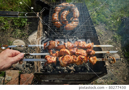Barbeque meat and sausages or bratwurst on a grill grate in backyard. Man preparing shashlik or shish kebab over charcoal. Grilled meat on metal skewer outdoor. BBQ party or picnic food. Close up shot 80277430