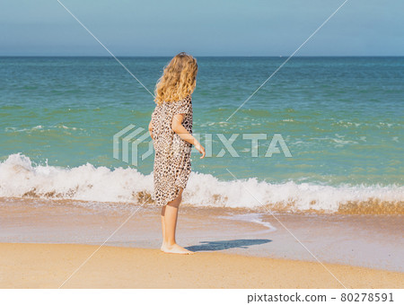 Young beautiful girl in beige dress staying along the sandy beach near the waves. Young beautiful girl in beige dress staying along the sandy beach near the waves. 80278591