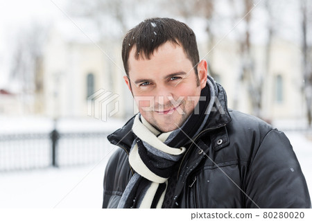 Young man in warm coat smiling at camera on blurred background of snowy park. Young man in warm coat smiling at camera on blurred background of snowy park. 80280020