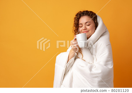 Sleepy young woman with curly hair covered in blanket and holding cup of coffee against yellow background Sleepy young woman with curly hair covered in blanket and holding cup of coffee against yellow background 80281120