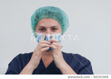 Close up of female doctor or scientist with a medical mask and surgical cap over grey background. She is adjusting mask with Close up of female doctor or scientist with a medical mask and surgical cap over grey background. She is adjusting mask with 80281908