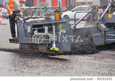 A road worker at the control panel of a tracked paver paver paving asphalt on a fenced road on a summer day. A road worker at the control panel of a tracked paver paver paving asphalt on a fenced road on a summer day. 80282081