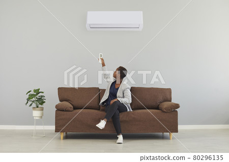 Black woman sitting on couch and switching her air conditioner with remote control 80296135