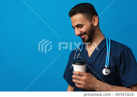 Young arab doctor drinking cup of coffee standing over blue background Young arab doctor drinking cup of coffee standing over blue background 80301828