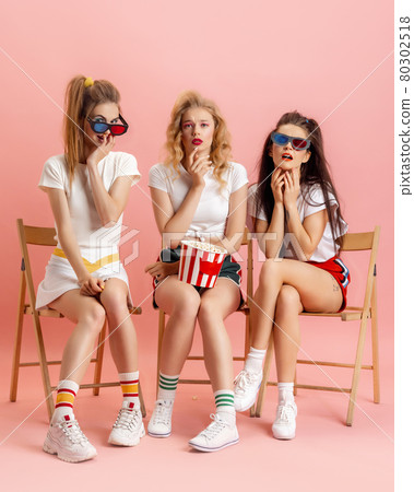 Three young women in retro 90s fashion style, outfits watching movie isolated over pink studio background. Concept of eras comparison, beauty, fashion and youth. 80302518
