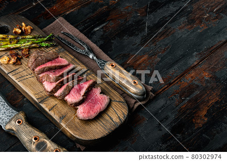 Sliced grilled marbled meat steak Filet Mignon, with onion and asparagus, on wooden serving board, with meat knife and fork, on old dark wooden table background, with copy space for text Sliced grilled marbled meat steak Filet Mignon, with onion and asparagus, on wooden serving board, with meat knife and fork, on old dark wooden table background, with copy space for text 80302974