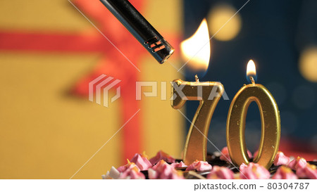 Birthday cake number 70 golden candles burning by lighter, background gift yellow box tied up with red ribbon. Close-up Birthday cake number 70 golden candles burning by lighter, background gift yellow box tied up with red ribbon. Close-up 80304787