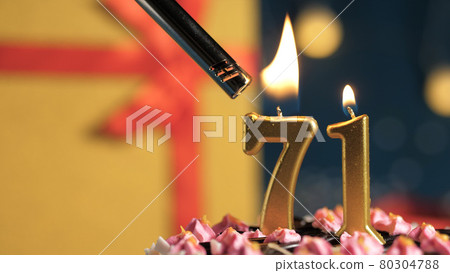 Birthday cake number 71 golden candles burning by lighter, background gift yellow box tied up with red ribbon. Close-up 80304788