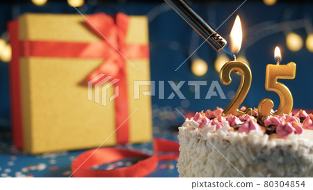 White birthday cake number 25 golden candles burning by lighter, blue background with lights and gift yellow box tied up with red ribbon. Close-up White birthday cake number 25 golden candles burning by lighter, blue background with lights and gift yellow box tied up with red ribbon. Close-up 80304854