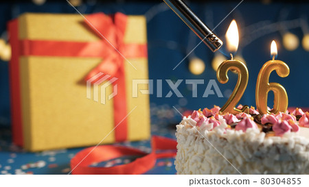 White birthday cake number 26 golden candles burning by lighter, blue background with lights and gift yellow box tied up with red ribbon. Close-up 80304855