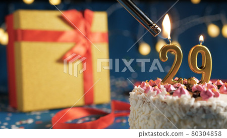 White birthday cake number 29 golden candles burning by lighter, blue background with lights and gift yellow box tied up with red ribbon. Close-up White birthday cake number 29 golden candles burning by lighter, blue background with lights and gift yellow box tied up with red ribbon. Close-up 80304858