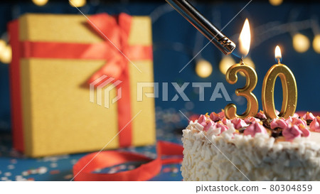 White birthday cake number 30 golden candles burning by lighter, blue background with lights and gift yellow box tied up with red ribbon. Close-up White birthday cake number 30 golden candles burning by lighter, blue background with lights and gift yellow box tied up with red ribbon. Close-up 80304859