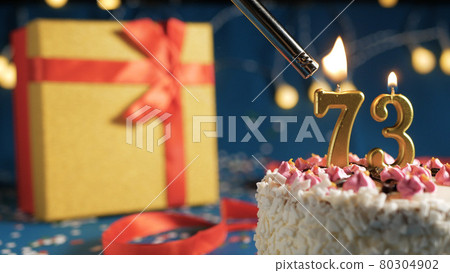 White birthday cake number 73 golden candles burning by lighter, blue background with lights and gift yellow box tied up with red ribbon. Close-up White birthday cake number 73 golden candles burning by lighter, blue background with lights and gift yellow box tied up with red ribbon. Close-up 80304902