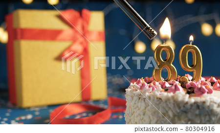 White birthday cake number 89 golden candles burning by lighter, blue background with lights and gift yellow box tied up with red ribbon. Close-up White birthday cake number 89 golden candles burning by lighter, blue background with lights and gift yellow box tied up with red ribbon. Close-up 80304916