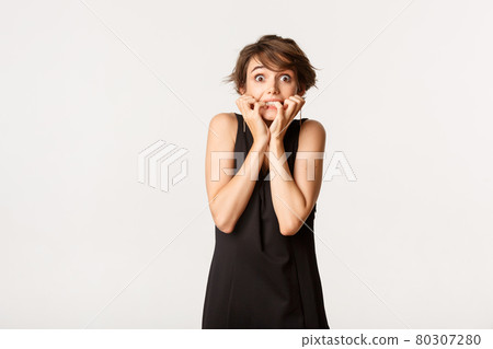 Image of scared and insecure brunette woman in black dress, trembling from fear and biting fingernails, standing over white background 80307280