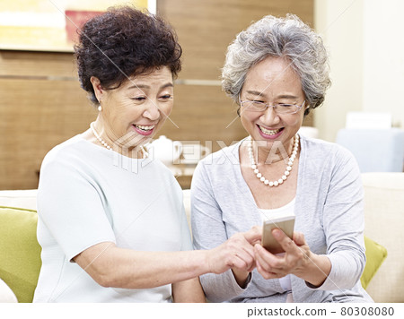 two senior asian women sitting on couch looking at cellphone, happy and smiling 80308080