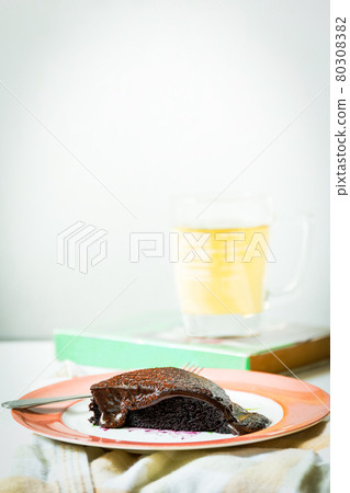 Delicious slice of moist chocolate cake on a plate with cup of tea in background. Delicious slice of moist chocolate cake on a plate with cup of tea in background. 80308382
