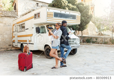Summer travel concept - Multiracial middle age couple or friends looking at map to plan a camper van road trip.African and latin ethnic. Holding big Travel bag or luggage . Trailer car on background. Summer travel concept - Multiracial middle age couple or friends looking at map to plan a camper van road trip.African and latin ethnic. Holding big Travel bag or luggage . Trailer car on background. 80309913