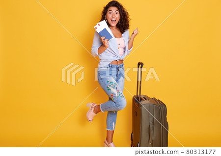 Funny happy girl posing on one leg near suitcase, holding passport with flight tickets, isolated over yellow background, clenching fists and looks happy. 80313177