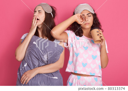 Closeup portrait of two sleeping female covering eyes and mouth, wearing pajamas and sleeping masks, girl with wavy hair drinking take away coffee, posing with closed eyes. Closeup portrait of two sleeping female covering eyes and mouth, wearing pajamas and sleeping masks, girl with wavy hair drinking take away coffee, posing with closed eyes. 80313374