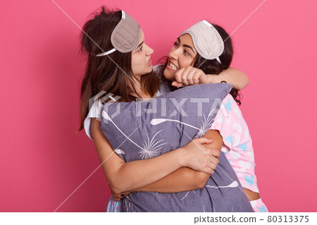 Two friends fighting during pajamas party, female embracing gray pillow, girl with grey blindfold choking her girlfriend, women standing against pink background. 80313375