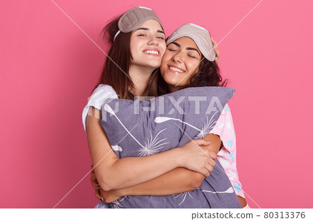 Two happy dark haired female wearing pajamas and blindfolds, hugging and embracing pillow, keeping eyes closed, expressing happiness, enjoying spending time together. Two happy dark haired female wearing pajamas and blindfolds, hugging and embracing pillow, keeping eyes closed, expressing happiness, enjoying spending time together. 80313376