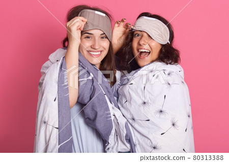 Two beautiful attractive friends laughing sincerely, having blindfold over foreheads, wearing pajamas, hugging, spending time together, ladies wrapped in blanket. Two beautiful attractive friends laughing sincerely, having blindfold over foreheads, wearing pajamas, hugging, spending time together, ladies wrapped in blanket. 80313388