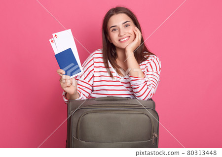 Smiling woman traveler holding passport with ticket posing with suitcase, looking directly at camera with happy facial expression, waiting her plane traveling abroad. 80313448