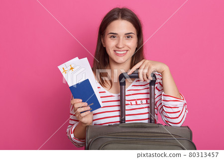 Close up portrait of happy European girl with suitcase, airplane tickets and passport isolated on rosy background, lady feels excited about trip, wearing casual white shirt with red stripes. Traveling Close up portrait of happy European girl with suitcase, airplane tickets and passport isolated on rosy background, lady feels excited about trip, wearing casual white shirt with red stripes. Traveling 80313457