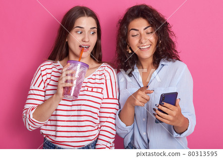 Picture of two good looking young women standing isolated over pink studio background, one lady holding cell phone and other holds water bottle and drinks, looking at device's screen with interst. Picture of two good looking young women standing isolated over pink studio background, one lady holding cell phone and other holds water bottle and drinks, looking at device's screen with interst. 80313595