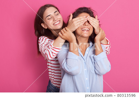 Close up portrait of beautiful Caucasian woman covering eyes to her friend standing behind and smiling, looking at girl wears blue casual shirt, posing isolated on pink background. Friendship concept. Close up portrait of beautiful Caucasian woman covering eyes to her friend standing behind and smiling, looking at girl wears blue casual shirt, posing isolated on pink background. Friendship concept. 80313596