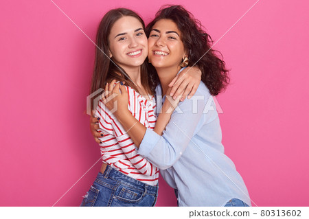 Indoor shot of two beautiful girls. One young woman hugs another, ladies looking smiling directly at camera, dresses casual shirts, brunette lesbian women isolated over pink. One sex love concept. 80313602