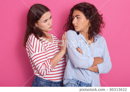Horizontal shot of young Caucasian female with dark hair puffing cheeks, pouting, having annoyed look, feeling displeased, looks at her girlfriend, another girl touches her shulder. Same sex love. 80313632